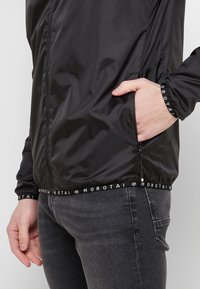 Veste légère noire avec fermeture éclair, en tissu texturé et poignets élastiques. Détails du logo sur la taille et les poches.