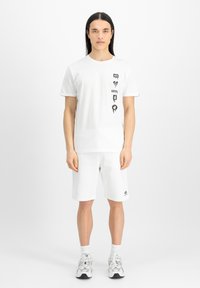 Tricou alb cu mânecă scurtă și grafica neagră, purtat cu pantaloni scurți albi. Îmbrăcămintea este dintr-un material confortabil, cu o croială lejeră.