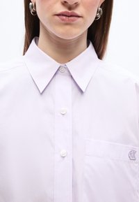 Γυναίκα που φοράει ένα ανοιχτό μωβ πουκάμισο με κουμπιά, με μυτερό γιακά και τσέπη στο στήθος που φέρει ένα μικρό κεντημένο λογότυπο.