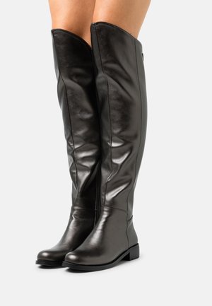 Vysoké černé kožené boty s nízkými podpatky nošené na bosých nohách na jednoduchém světlém pozadí.