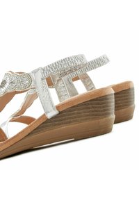 Sandali a cuneo argentati con una fascia texturizzata, impreziosita da perline. Sottopiede beige e suola in gomma marrone con scanalature verticali.