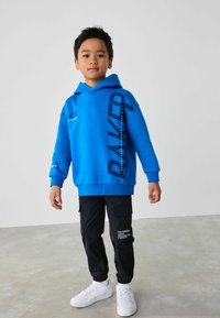Blauwe hoodie met logografieken, gecombineerd met zwarte cargobroeken met zakken en witte sneakers. Gladde stof en casual ontwerp.