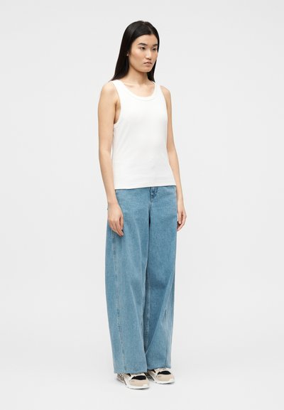 Γυναίκα που στέκεται με το σώμα της ελαφρώς στραμμένο προς τα δεξιά, φορώντας άσπρο μπλουζάκι χωρίς μανίκια, φαρδιά μπλε τζιν παντελόνια και μπεζ με μαύρα αθλητικά παπούτσια, μπροστά σε απλό φόντο.