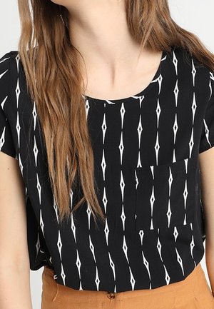 Mujer que lleva una blusa negra con patrones verticales de diamantes blancos y un bolsillo en el pecho, combinada con pantalones de talle alto de color mostaza.