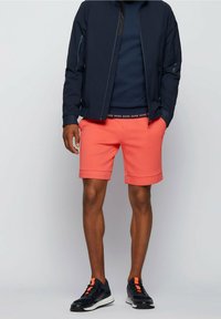 Uomo che indossa pantaloni corti corallo con una cintura con logo nero, giacca blu e sneakers nere con strisce arancioni, in piedi contro uno sfondo chiaro e neutro.