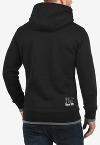 Zwarte hoodie met een gladde textuur, uitgevoerd met een contrasterende grijze zoom en logodetail ter hoogte van de taille, getoond vanuit de achterkant.