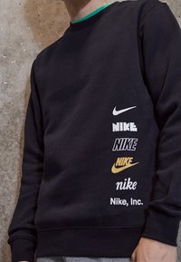 Person med svart sweatshirt som har sex vertikala Nike-logotyper i olika typsnitt och färger på högra sidan mot en texturerad väggbakgrund.