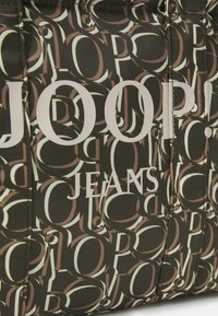 Hnědá vzorovaná látka s bílým textem "JOOP! JEANS" a překrývajícími se kruhovými vzory v různých odstínech. Texturovaný povrch.