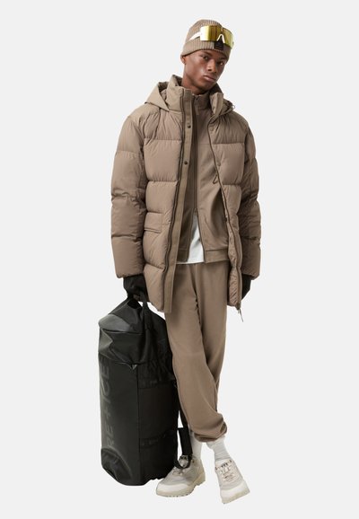 Puffer jacket in light brown with a hood, paired with a matching tracksuit. Black duffel bag and beige sneakers complete the look.