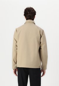 Beige, hosszú ujjú dzseki egyenes szabással és galléros nyakkal, finom varrási részletekkel, sima textúrával és patentos záródással az alján.