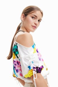 Hvit bluse med fargerike blomster mønstre i lilla, gul og turkis. Har av- skulder design og elastiske rynkede ermer.