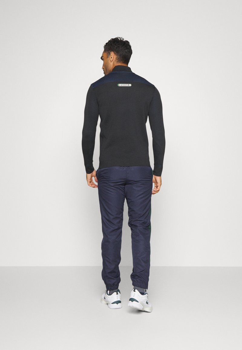 Homme debout de dos, portant une chemise noire à manches longues avec le logo Lacoste dans le dos, un pantalon marine et des baskets blanches sur un fond clair.