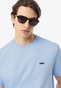 Lichtblauwe katoenen t-shirt met een ronde hals en korte mouwen; heeft een gekleurd krokodil logo op de borst. Donkere zonnebril met een rechthoekig montuur.