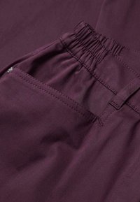 Pantalons bordeaux avec une texture lisse, dotés d'une taille élastique et d'une poche latérale avec des détails de couture visibles.