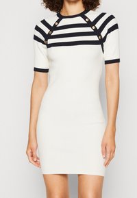 Robe blanche côtelée avec des rayures bleu marine, manches courtes et boutons décoratifs dorés aux épaules. Design ajusté avec une texture lisse.