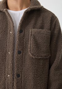 Jachetă din fleece maro cu o finisare texturată, nasturi cu împingere și o mare buzunar pe partea stângă. Culoare și deschidere frontală vizibile.