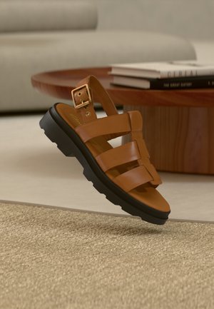 Καφέ δερμάτινο σανδάλι με μαύρη χοντρή σόλα και χρυσή αγκράφα στο λουράκι, αιωρούμενο πάνω από μπεζ υφασμένο χαλί κοντά σε στρογγυλό ξύλινο τραπέζι και βιβλία.