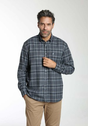 Homme aux cheveux courts et foncés portant une chemise à carreaux grise et bleue à boutons et un pantalon beige, debout devant un fond blanc.