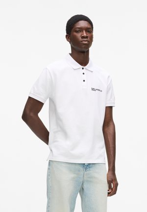 Joven con una camisa polo blanca de manga corta con botones negros y unos jeans azul claro, sobre un fondo blanco liso.