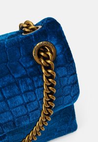 Modrý sametový kabelka s krokodýlí texturou, se zlatým řetízkovým držadlem a označením značky na boku.