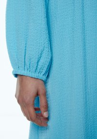 Hellblaues, strukturiertes Kleid mit einem gathered Ärmelbund, der die Weichheit des Stoffes und das zarte Muster zeigt und dabei die Handplatzierung auf dem Kleidungsstück hervorhebt.