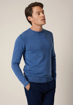 Mężczyzna ubrany w dopasowany niebieski sweter z okrągłym dekoltem i ciemne spodnie, stojący z rękami w kieszeniach, spoglądający lekko na bok.