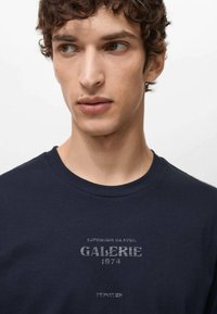 Navy blue cotton t-shirt with round neckline. Features a distressed graphic print "GALERIE 1974" in grey on the front. Smooth texture.