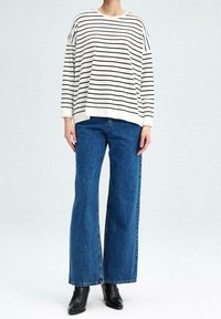 Person in einem weißen Langarmshirt mit schwarzen horizontalen Streifen, blauen weiten Jeans und schwarzen Knöchelstiefeln, steht vor einem weißen Hintergrund.