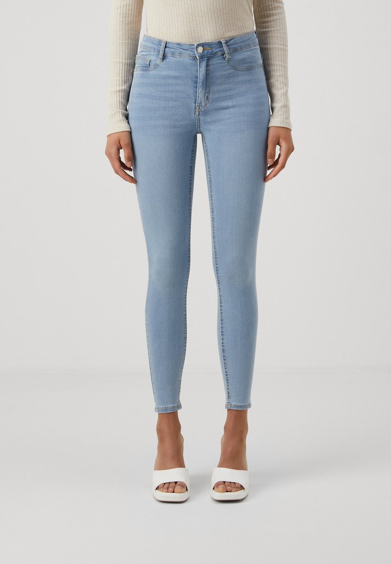 Jeans skinny de cintura alta em azul claro, com uma textura suave e ligeiramente desbotada. Apresenta bolsos frontais, um botão e um fecho com zíper.