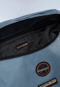 Borsa di stoffa blu con interni neri, dotata di una toppa e logo all'esterno. Chiusura con zip sulla parte superiore, texture liscia e design semplice.