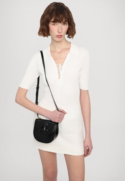 Witte geribbelde jurk met een kraag, korte mouwen en een vetersluiting bij de hals, gecombineerd met een kleine zwarte crossbody tas met een unieke sluiting.
