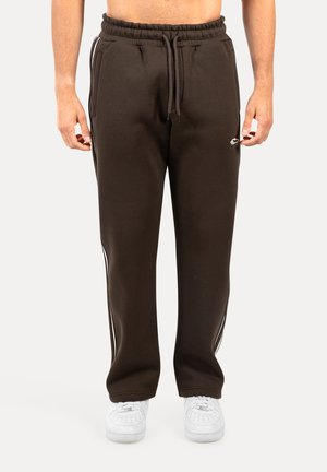 Brązowe dresy z miękkiego materiału, z białymi bocznymi paskami, elastycznym pasem i przednimi kieszeniami. Noszone z białymi sneakersami.