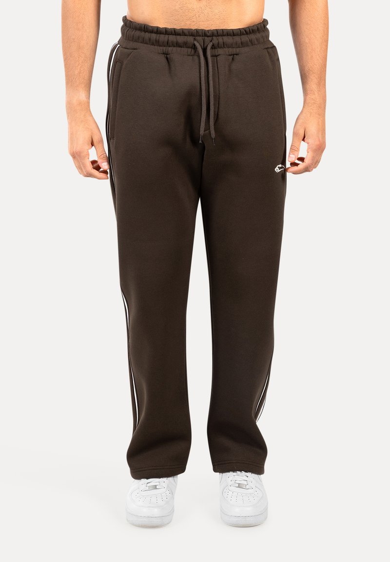 Brązowe dresy z miękkiego materiału, z białymi bocznymi paskami, elastycznym pasem i przednimi kieszeniami. Noszone z białymi sneakersami.