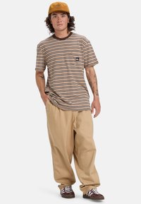Camisa de manga corta a rayas en marrón y beige con un bolsillo en el pecho, combinada con pantalones holgados beige y zapatillas marrones, rematada con una gorra marrón.