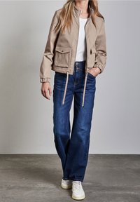 Veste beige avec col montant, fermeture éclair et deux poches avant ; associée à un jean bleu et des baskets blanches sur un fond neutre.