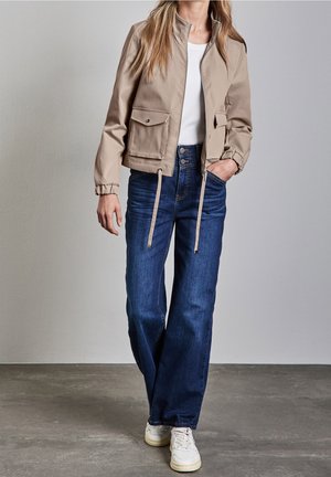 Veste beige avec col montant, fermeture éclair et deux poches avant ; associée à un jean bleu et des baskets blanches sur un fond neutre.