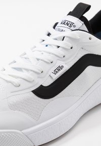Baskets blanches Vans avec détail bande noire, lacets blancs, tissu en mesh et étiquettes de marque sur la languette et le panneau latéral.