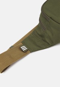 Grön tygaxelväska med ett tanfärgat band, som har en dragkedjestängning, texturerad yta och en liten etikett med tre vita ränder.