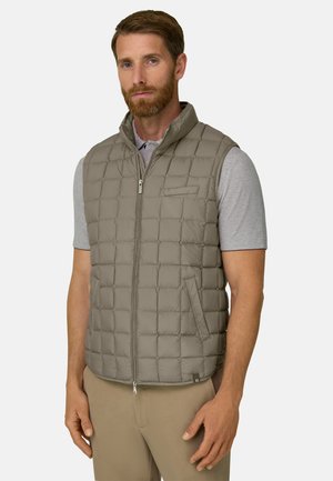 Homme barbu portant une chemise gris clair à manches courtes et un gilet matelassé beige zippé devant un fond uni.