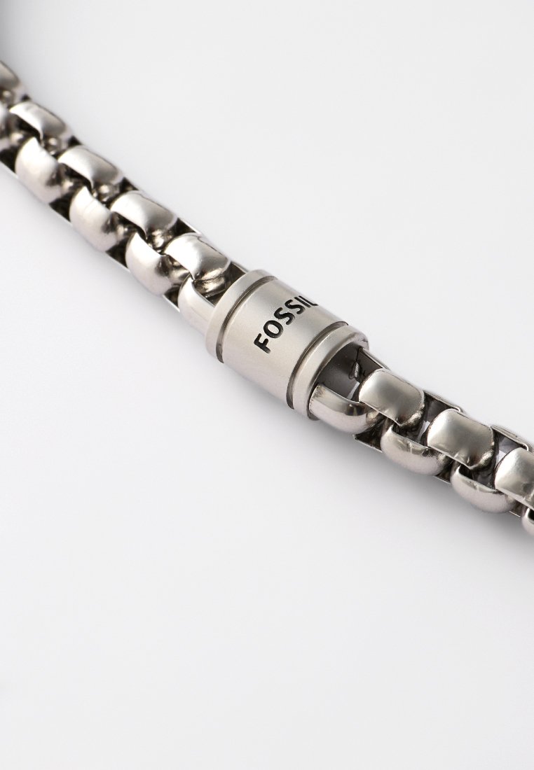 Bracelet en chaîne métallique argentée avec un fermoir cylindrique gravé du nom de la marque "FOSSIL" sur un fond blanc.