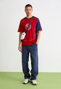 Jersey rojo y azul con acentos amarillos, combinado con pantalones oscuros holgados. Bolsa cruzada blanca y zapatillas gruesas blancas. Texturas suaves.