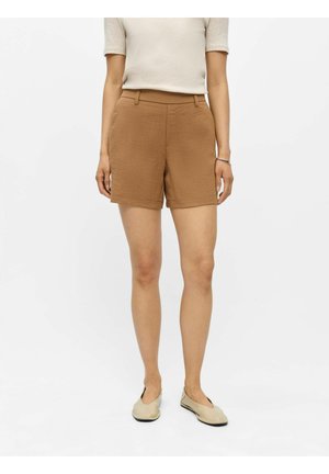 Kvinde står iført beige kortærmet top, brune teksturerede højtaljede shorts, beige flade sko og et armbånd på venstre håndled.
