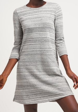 Frau trägt ein knielanges, langärmliges, grau-weiß gestreiftes Freizeitkleid mit rundem Ausschnitt vor einem schlichten Hintergrund.