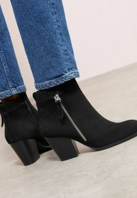 Botines de ante negros con tacón cuadrado, que presentan un cierre lateral con cremallera y un detalle de borlas. Combinados con jeans de mezclilla azul.