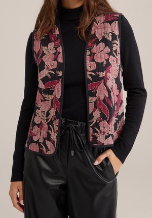 Vrouw die een zwarte top met lange mouwen draagt, gecombineerd met een zwart-roze gebloemd doorgestikt vest en zwarte broek van nepleer met een trekkoord in de taille.