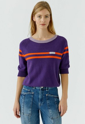 Femme portant un pull violet avec deux bandes horizontales orange et un jean bleu avec de grandes poches avant, debout devant un fond uni.