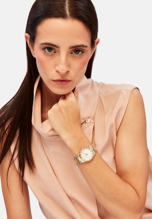Montre en métal doré et argenté au poignet, avec un cadran blanc rond, des marqueurs d'heure minimalistes et une finition polie. Bords arrondis et design élégant.