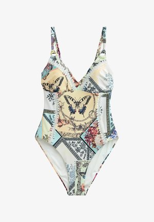 Femme debout à l'extérieur portant un maillot de bain une pièce à motifs papillons et floraux, sur fond de ciel bleu et rochers.