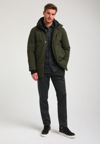 Giacca impermeabile verde oliva con cappuccio, abbinata a una camicia scura dalla texture e pantaloni neri a fantasia, e sneakers nere con suola bianca.