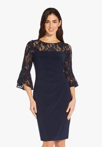 Žena se usmívá a má na sobě tmavě modré šaty po kolena s krajkovými rukávy a průkrčníkem a s nařaseným detailem látky v pase.